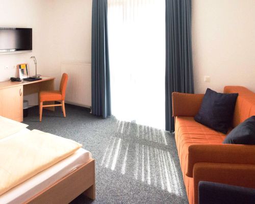 Standard-Dreibettzimmer im Johanniter Gästehaus Münster mit Bett, Schreibtisch, Sofa, TV und großem Fenster.