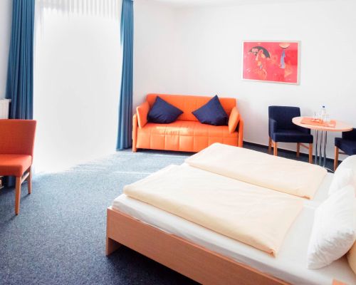 Standard-Dreibettzimmer mit Doppelbett, orangefarbenem Sofa, kleinem Tisch und Fensterfront.