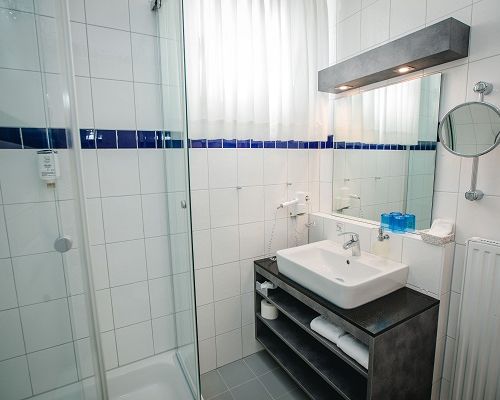 Badezimmer zum Standard-Dreibettzimmer mit Waschbecken, Dusche, Glasabtrennung und weißen Fliesen mit blauem Akzent.